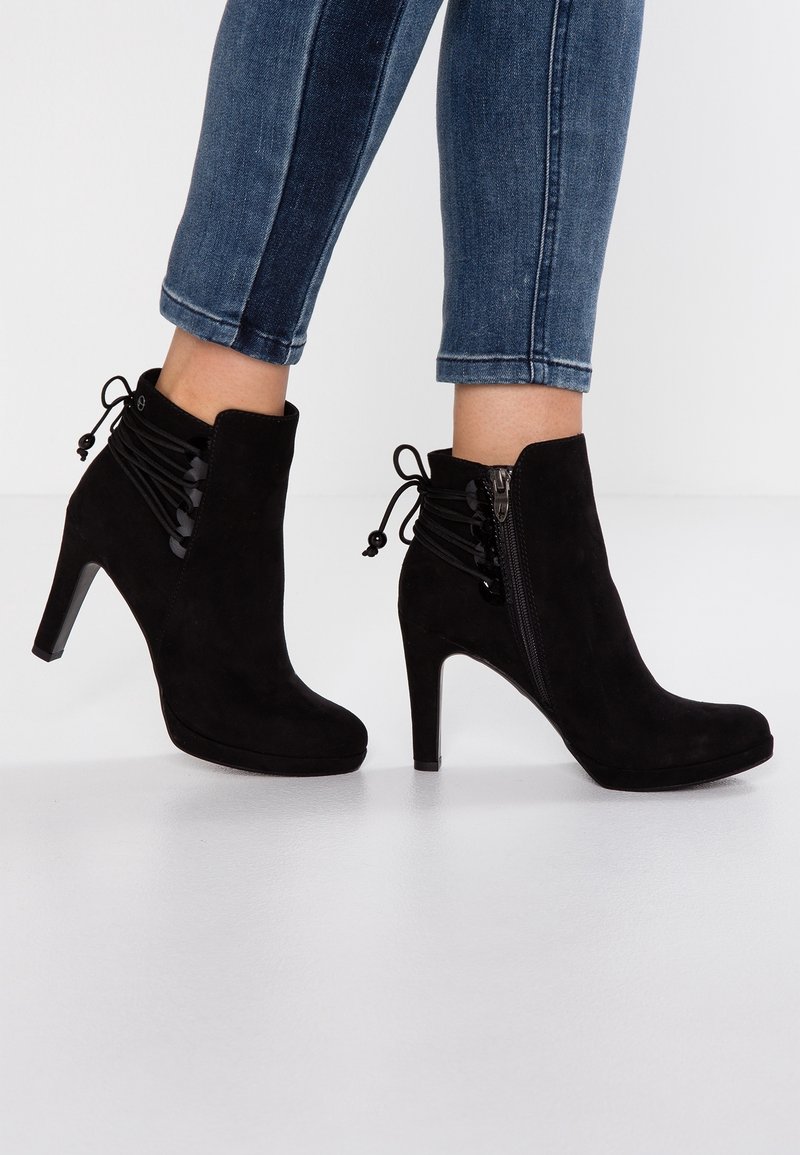Bottes cheville en daim noir avec talons hauts, fermeture éclair latérale et lacets décoratifs, portées par une personne en jean skinny bleu.