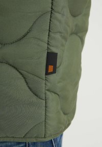 Veste matelassée verte avec une finition texturée, dotée d'une étiquette noire avec un accent orange, des coutures visibles et un ourlet ajusté.