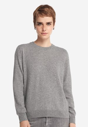 Młoda osoba z krótkimi włosami ubrana w jednolity, szary sweter z okrągłym dekoltem oraz pasujące szare spodnie na białym tle.