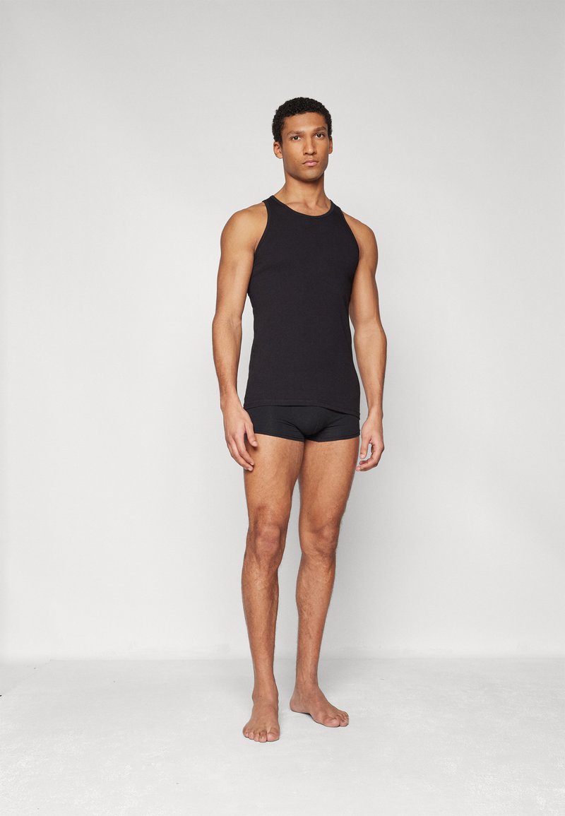 Czarny top na ramiączkach wykonany z bawełny, z okrągłym dekoltem i bez rękawów. Noszony z czarną dopasowaną bielizną i boso.