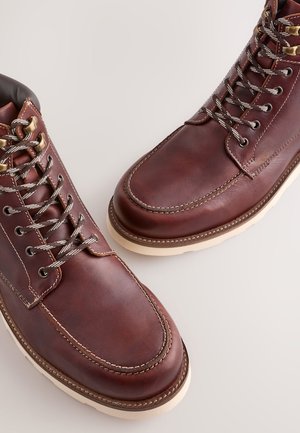 Bruine leren laarzen met een gladde afwerking, voorzien van metalen veteroogjes, grijze veters en een rubberen zool in crèmekleur. Stiksel detail langs de randen.