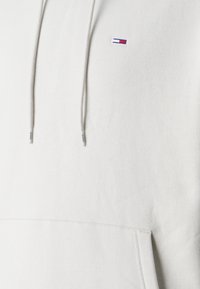 Ljust grå huvtröja med frontficka, dragsko och en liten, färgglad logga på vänster bröst. Slät bomullsblandning.