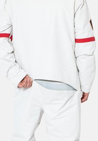 Prenda exterior blanca con una franja roja en el brazo. Cremallera trasera parcialmente abierta, revelando una tela interior en un color contrastante.