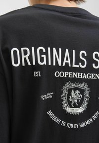 Schwarzes Baumwoll-T-Shirt mit weißem Text und einem grafischen Design mit einem Wappen, das ein Pferd und dekorative Elemente umfasst.