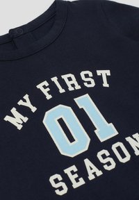 Tmavě modré bavlněné tričko s kulatým výstřihem, s potiskem textu "MOJE PRVNÍ 01 SEZÓNA" v bílé a světle modré barvě na přední straně.