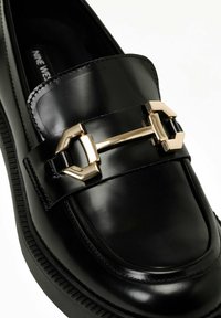 Mocassin en cuir noir avec un accent en métal doré design. Texture lisse, bout arrondi et semelle en caoutchouc noir avec des détails en relief.