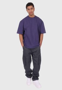 Camiseta morada de gran tamaño, pantalones cargo gris holgados con bolsillos laterales y zapatillas blancas, usados por una persona que está de pie frente a un fondo sencillo.