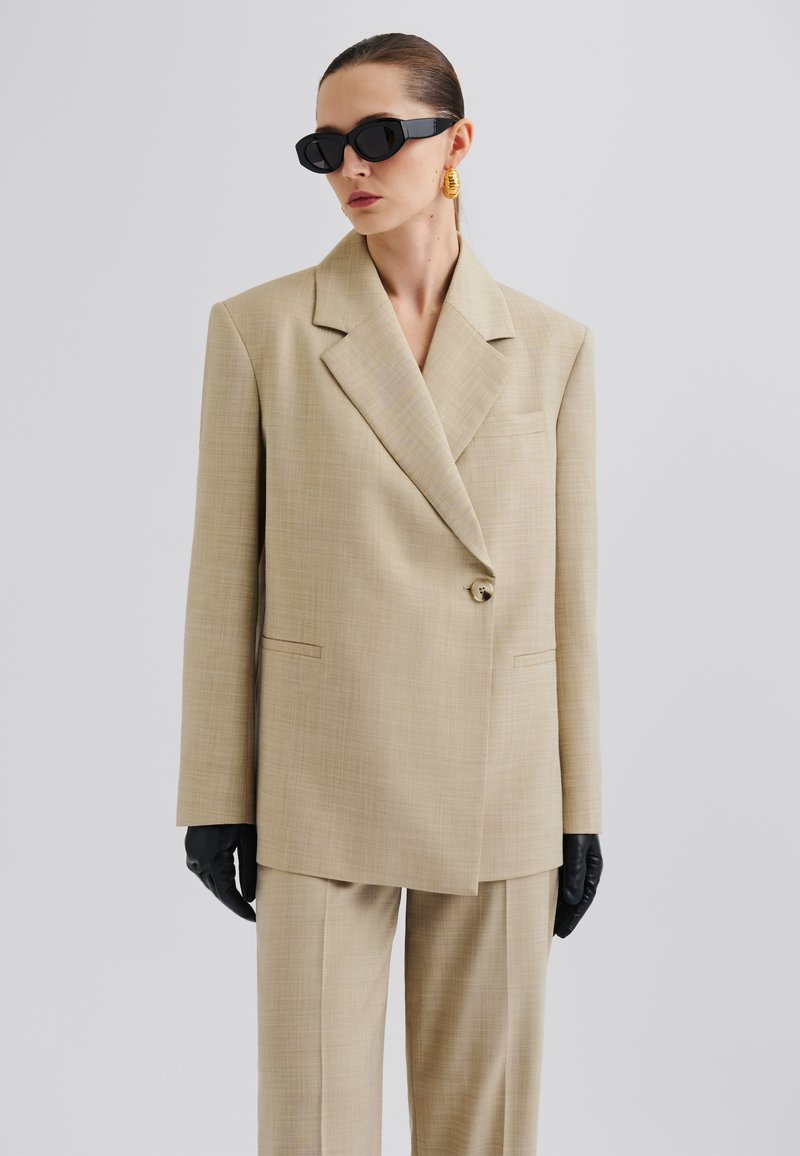 Beige tailored blazer with a notch lapel, single button closure, and two front pockets, paired with matching trousers and black gloves.