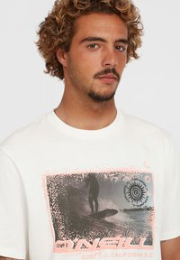 Valkoinen puuvillapaita, jossa on graafinen printti surffaajasta, ja kuvan ympärillä on vaaleanpunaisia ja mustia yksityiskohtia. Pyöreä pääntie.
