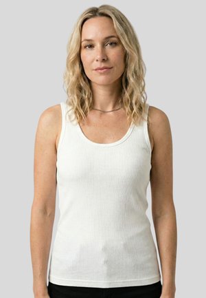Lyshåret kvinde med bølget hår iført en hvid ribbet tanktop og en tynd halskæde, stående foran en ensfarvet grå baggrund.