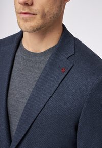 Granatowa marynarka z teksturowanej tkaniny, z wyrazistym kołnierzem i małym czerwonym emblematem na klapie, nałożona na szary shirt.