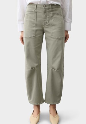 Vooraanzicht van een persoon die khaki wijde, hoog getailleerde broek draagt met grote voorzakken en beige instapschoenen.