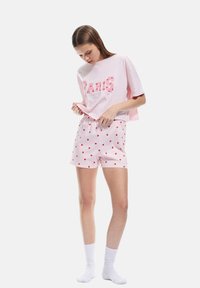 Rosa Baumwoll-T-Shirt mit dem Schriftzug "PARIS", kombiniert mit weißen Shorts, die rote Herzmuster und pinke Streifen aufweisen. Weiße Socken runden das Outfit ab.