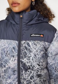 Femme portant une doudoune marbrée gris et blanc avec un col montant, une capuche, une fermeture éclair à l'avant, et un patch logo ellesse sur la poitrine.
