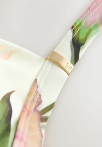 Primo piano della cinghia in tessuto stampato floreale con cuciture bianche e anello metallico regolabile color oro su uno sfondo chiaro.