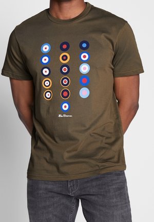 Voksen iført olivengrøn T-shirt med tre lodrette kolonner af farverige cirkulære måldesigns i rød, blå, gul og hvid på brystet.