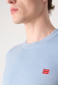 Hellblaues Baumwollcrewneck-T-Shirt mit einem kleinen roten rechteckigen Etikett mit "HUGO" in schwarzem Text auf der linken Brustseite.