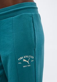 Φόρμες Teal Puma Athletics με πλαϊνή τσέπη και λευκή λογότυπη επιγραφή "Puma Athletics Since 1948" στο αριστερό μηρό, σε λευκό φόντο.
