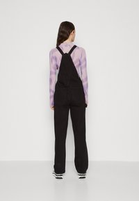 Black denim overalls with adjustable straps and pockets, paired with a long-sleeve purple striped top. Sneakers feature black and white accents.