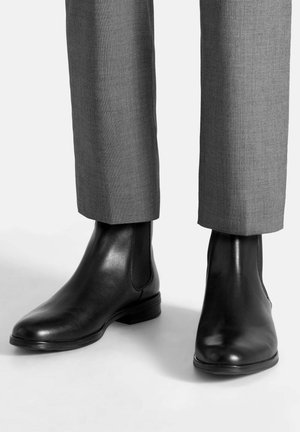 Czarne skórzane botki za kostkę z elastycznymi panelami po bokach, gładką fakturą, lekko spiczastym noskiem, prezentowane na tle szarych, dopasowanych spodni.