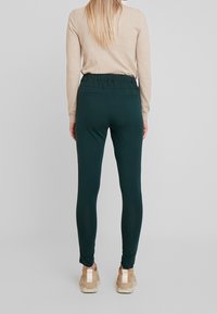 Legging vert, extensible avec une taille élastique, présentant une texture lisse et deux poches arrière. Porté avec un haut à manches longues beige et des baskets.