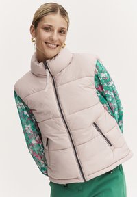 Vesta căptușită de culoare roz pal, cu guler înalt, fermoar dublu și două buzunare laterale. Mânecele au un imprimeu floral multicolor pe un fond verde.