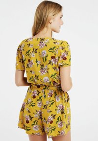 Combinaison florale en tissu jaune avec des manches courtes. Présente des fleurs violettes et blanches, des feuilles vertes, une taille élastique et des poches latérales.