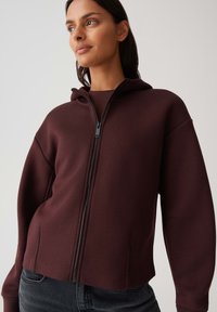Bordeaux rits hoodie met een gestructureerd ontwerp, zachte textuur en zijsplitten; voorzien van een minimale zwarte rits en ribgebreide manchetten.
