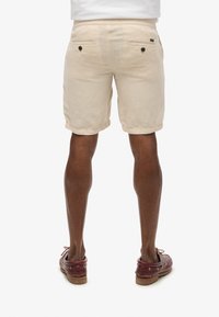 Pantalones cortos de lino beige con dobladillos, dos bolsillos traseros con botones oscuros, usados con zapatos de cuero marrón. Vistos desde atrás.