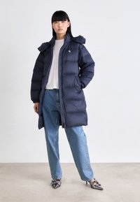 Marinblå puffjacka med dragkedja, avtagbar huva, horisontell quiltning och en liten logotyp. Kombinerad med ljusblå jeans och silverfärgade klackar.