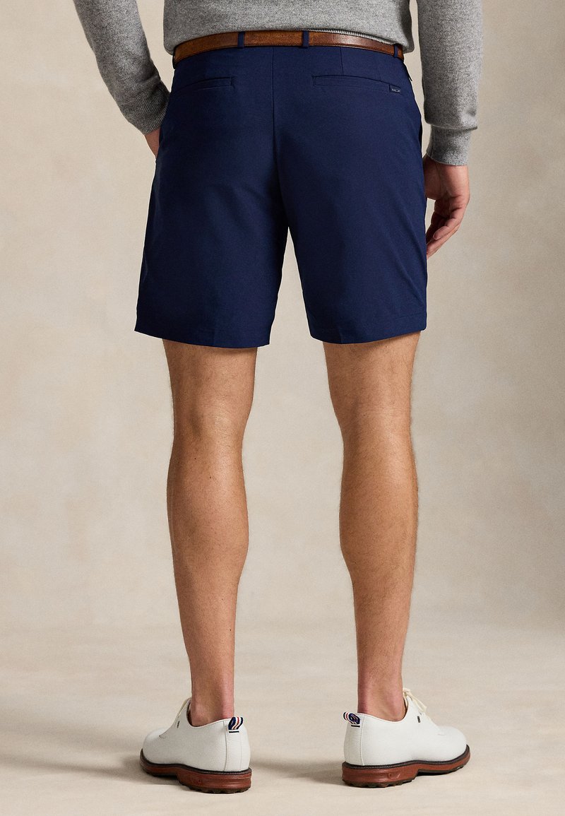 Marineblaue Shorts aus leichtem Stoff mit glatter Textur, zwei Gesäßtaschen und einem braunen Gürtel. Weiße Schuhe vervollständigen den Look.