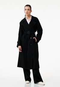 Abrigo negro de trench en una tela fluida con cintura ceñida, gran solapa, botones de doble pecho y bolsillos laterales. Se lleva sobre pantalones de pernera ancha.
