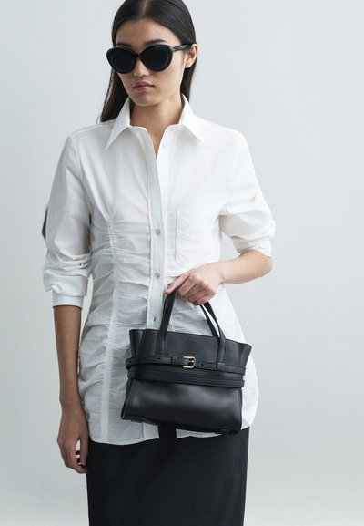 Γυναίκα που φορά μαύρα γυαλιά ηλίου και λευκό πουκάμισο με πτυχώσεις κρατά μια μαύρη δερμάτινη τσάντα με λεπτομέρεια από αγκράφα μπροστά σε ένα απλό φόντο.