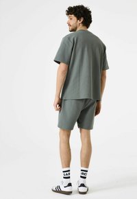 Heren twee-delig outfit in gestructureerde grijze stof, bestaande uit een losse top met korte mouwen en bijpassende shorts, gecombineerd met witte sokken en sneakers.