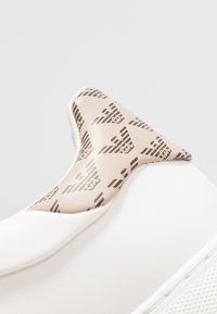 Vit sko med beige vadderad tunga som har upprepande mörka örnlogotyper, närbild som visar texturerad sula och slätt lädermaterial.