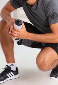 Šedé sportovní tričko, černé šortky s barevnými akcenty, drží průhlednou lahev na vodu. Černé tenisky s bílými pruhy. Čisté pozadí.