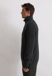 Pull gris foncé avec une texture côtelée et un col haut. Présente des fentes latérales et des manches longues. Fabriqué en matière tricotée douce.