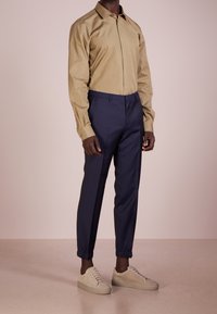 Långärmad skjorta i tan, marinblå tapered-byxor och beige mockasneakers. Skjortan har en klassisk krage; byxorna har elastiska muddar och rena linjer.