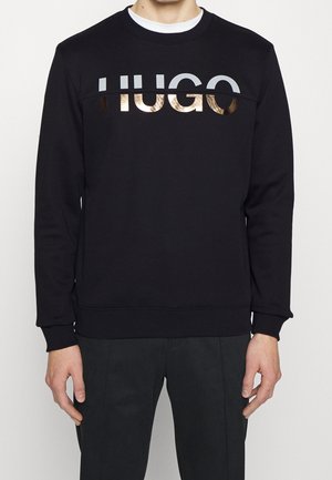 Mężczyzna noszący czarną bluzę z białym i metalicznym złotym logo "HUGO" na wysokości klatki piersiowej, zestawioną z czarnymi spodniami.