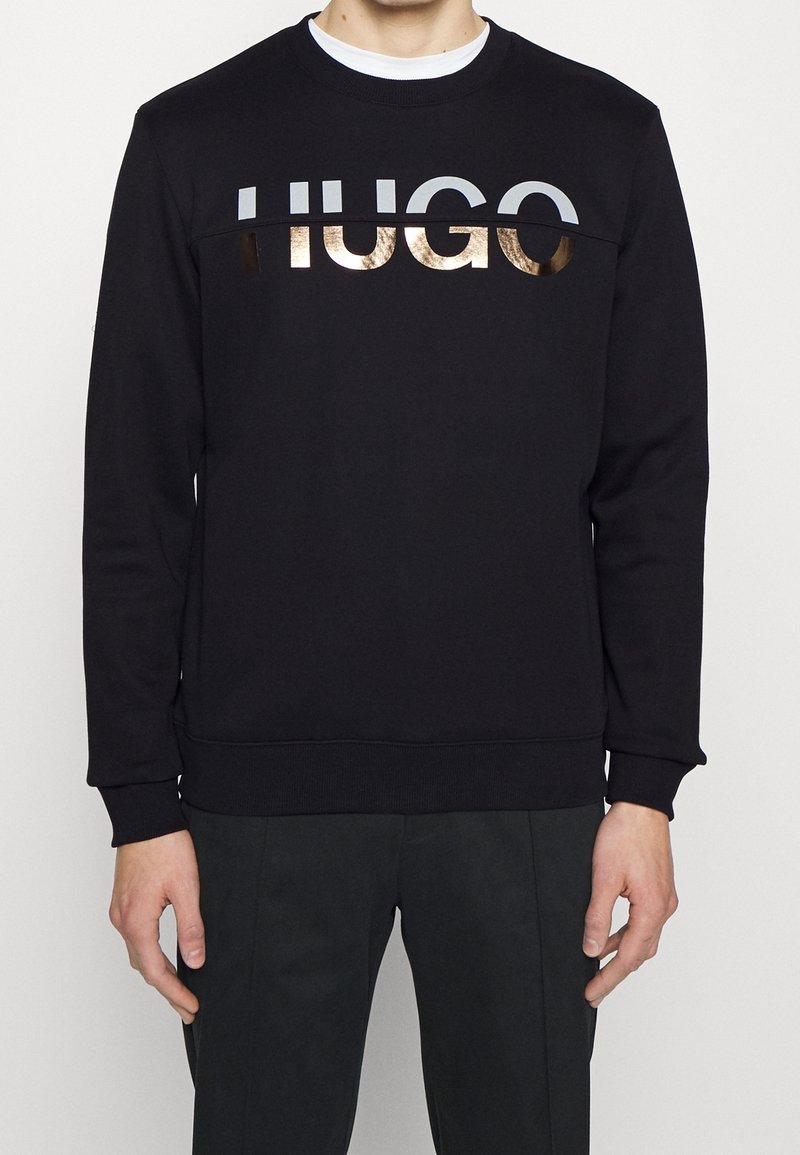 Homme portant un sweat-shirt noir avec un logo "HUGO" blanc et doré métallique sur la poitrine, associé à un pantalon noir.