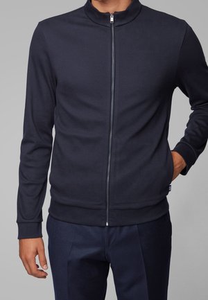 Mężczyzna ubrany w granatową teksturowaną kurtkę na zamek z długimi rękawami oraz dopasowane granatowe spodnie, z jedną ręką w kieszeni.