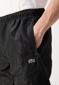 Černé, lehké kraťasy s texturovaným povrchem, elastickým pasem, bočními kapsami a zeleným logem krokodýla Lacoste na levé straně.