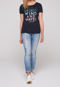 Tmavě modré pruhované tričko s barevným nápisem "DUCH VĚTRU A VLN". Světle modré džíny s bílými textovými akcenty. Bílé tenisky doplňují outfit.