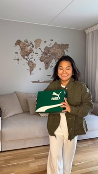 Puma-skorbox med grön utsida, vitt logotyp och kurvigt design. En person håller i den, klädd i en olivgrön jacka och vita byxor.