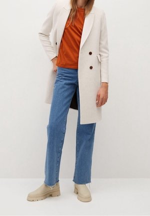 Personne portant un manteau beige à double boutonnage, un haut couleur rouille, un jean bleu droit et des bottes beiges épaisses, debout devant un fond blanc uni.