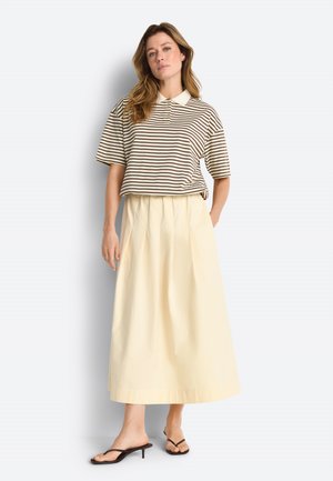 Kobieta o falowanych włosach, ubrana w brązowo-białą koszulkę polo w paski, kremową długą spódnicę i czarne sandały na obcasie, stojąca na białym tle.