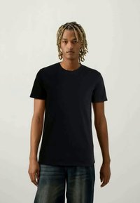 Mlad moški z blond dreadi, oblečen v navadno črno majico in zbledela modra kavbojka, stoji ob nevtralni ozadju.