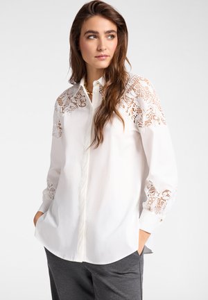 Femme portant un chemisier blanc avec des détails en dentelle florale sur les épaules et les manches, associé à un pantalon gris foncé, les mains dans les poches, regardant vers la droite.