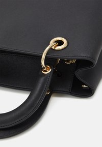 Czarna torebka strukturalna z teksturowanym wykończeniem, z złotym metalowym pierścieniem i elementami, oraz krzywym czarnym uchwytem.