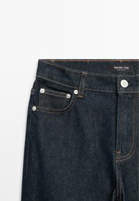 Jeans in denim scuro con cuciture arancioni, dotati di tasca frontale, chiusura con bottone e etichetta del marchio in vita. Texture liscia.